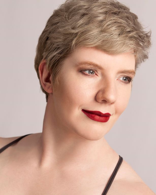 Laurel, a white woman with creamy skin, cropped silvery hair, and hazel eyes, looks thoughtfully to one side.  Crimson lipstick punctuates a slight smile.